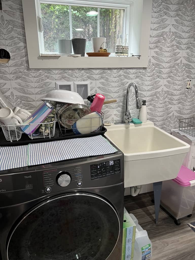 Laundry room sink used to do dishes; dishes drying on a rack on top of the clothes dryer. 