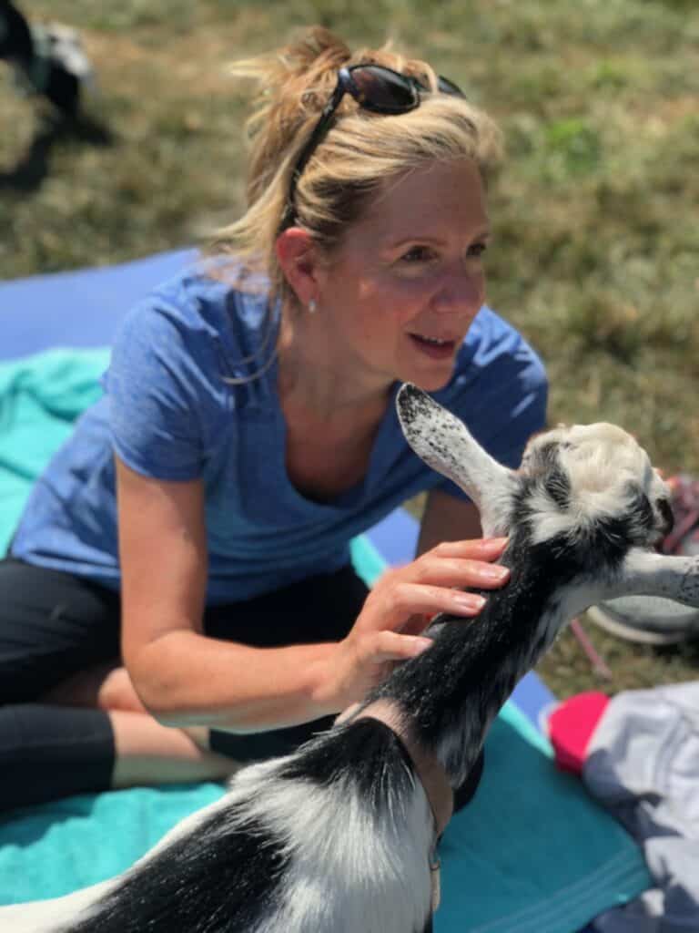 A woman sitting on a yoga mat petting a goat.