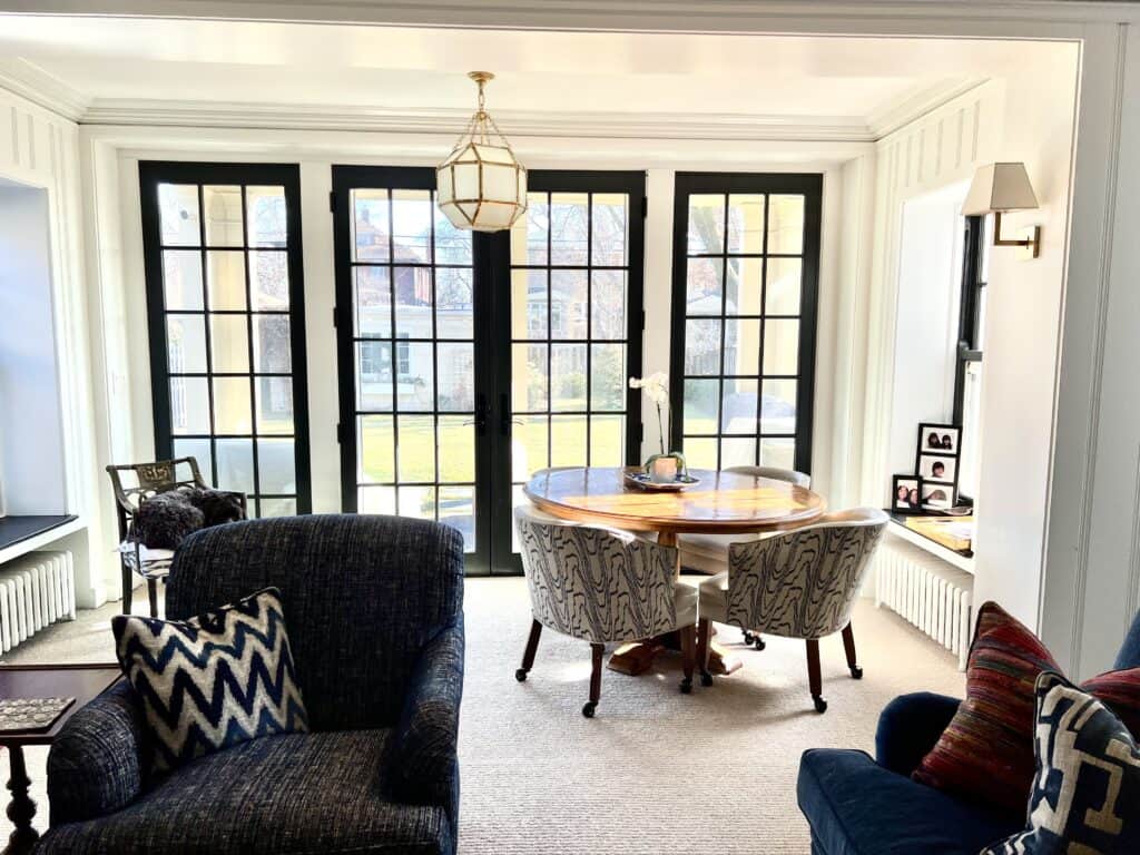 Photo of the family room with chair and sofa in the foreground and game table and chairs by the wall of windows.