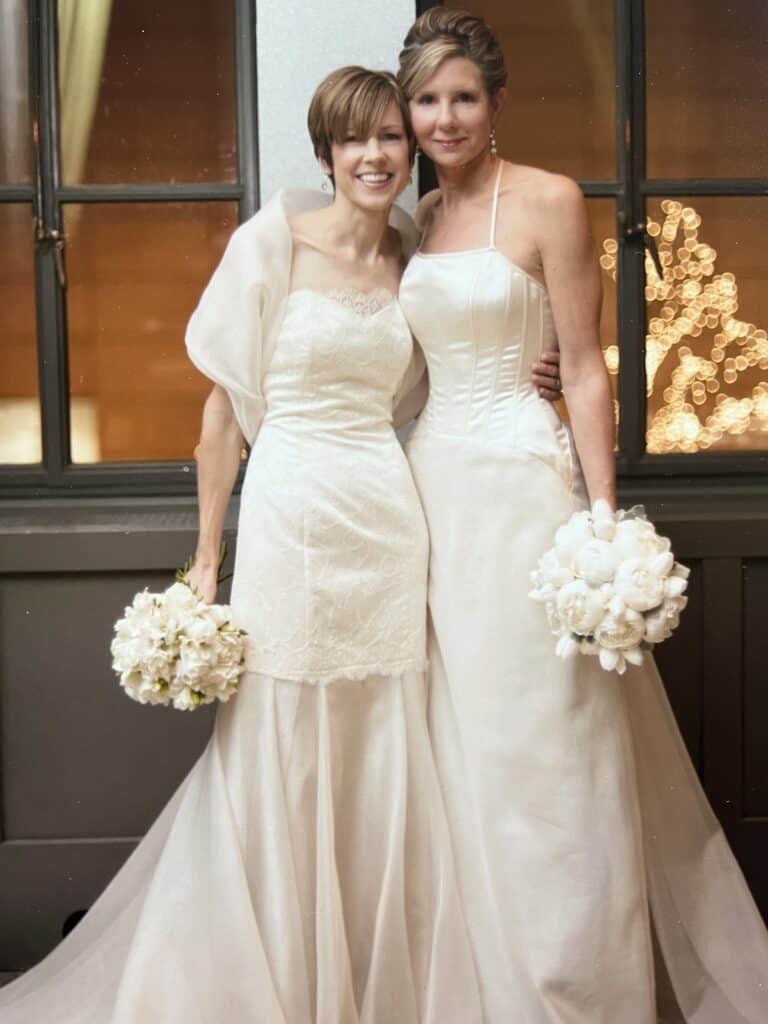 Two women in beautiful wedding gowns surrounded by a window with sparkly lights beyond. 