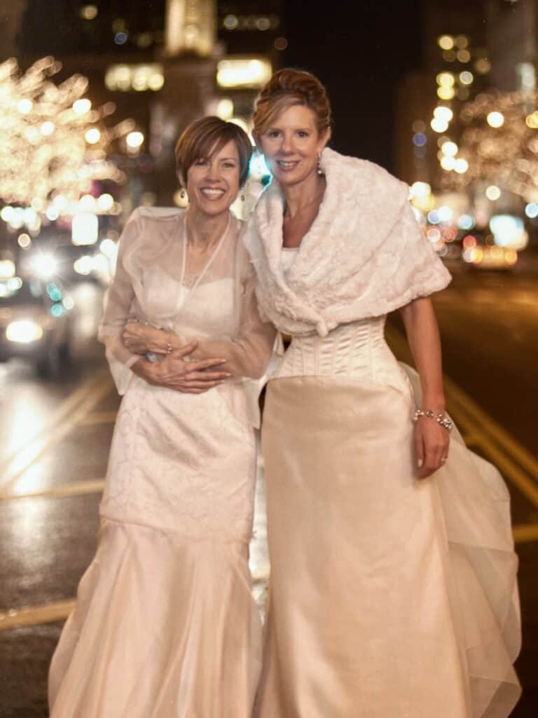 Two women in wedding dresses standing in the middle of Michigan Avenue in Chicago, surrounded by cars and holiday lights. 