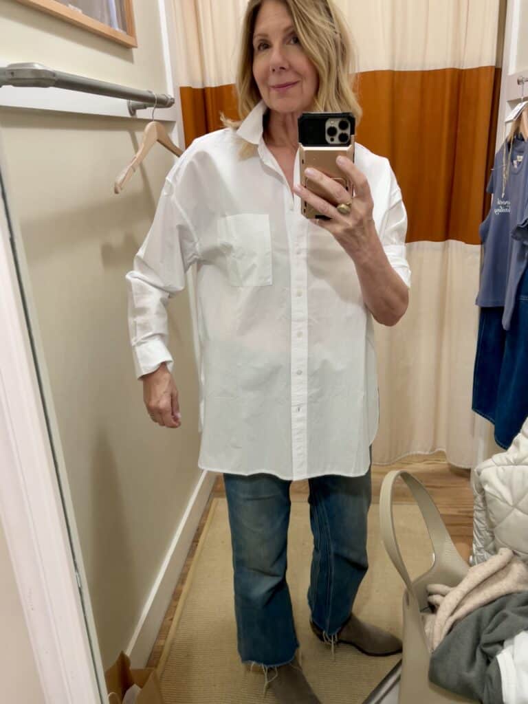 Woman in a dressing room trying on an oversized white button down shirt. 