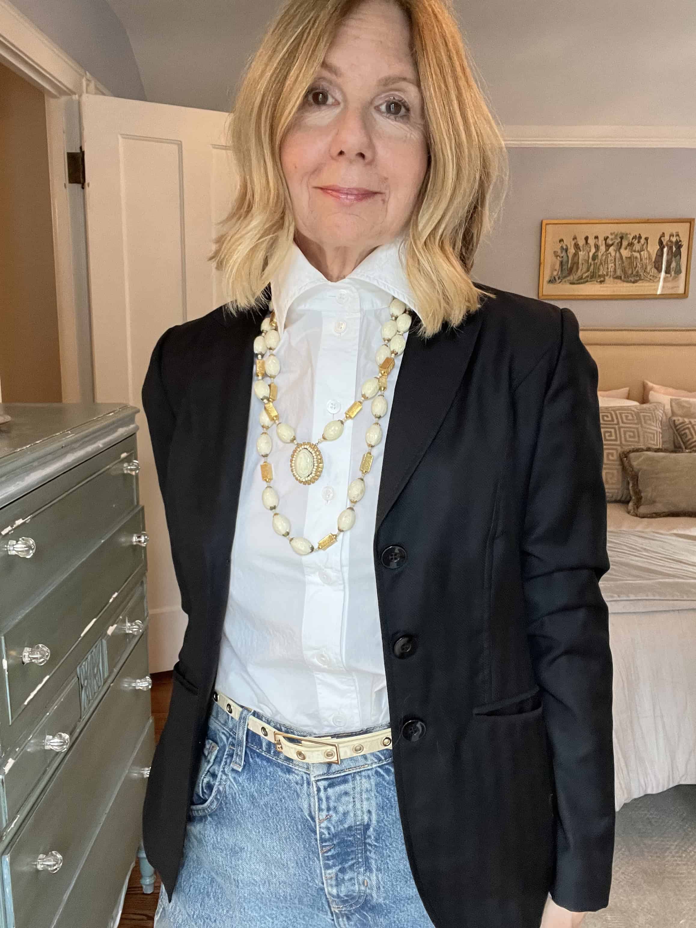 A woman styling a white button down shirt, buttoned at the collar, with chunky necklaces and a blazer. 