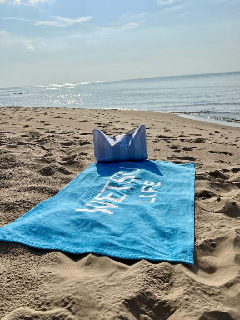 The teal WETSU Life beach towel and beach bag laying on the sand by the lake. 