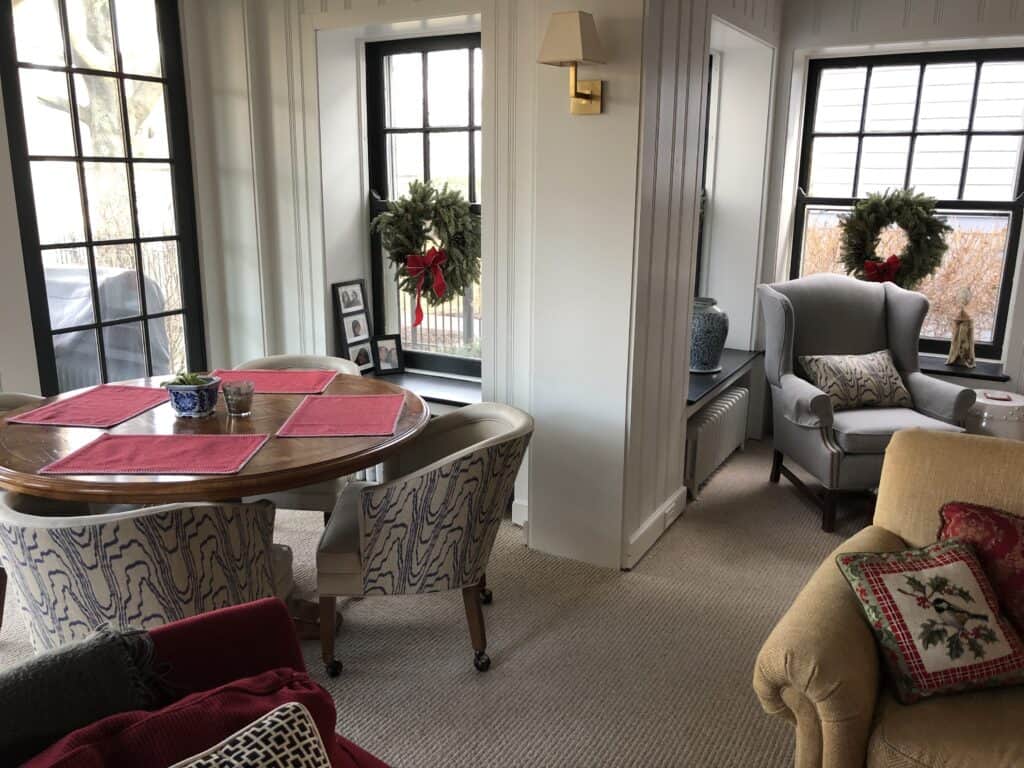 Corner of a room adorned with simple Christmas decor including wreaths, red placemats and a needlepoint pillow. 