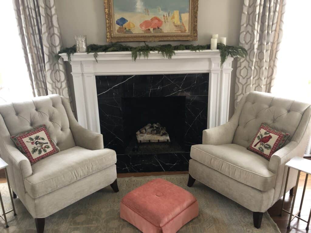 Photo of a fireplace mantle draped with cedar garland, surrounded by two chairs with festive needlepoint pillows. 